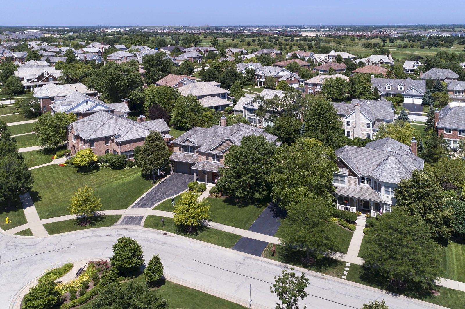 Placeholder. Aerial view of a suburban street of two-story homes with shingle roofs and mature trees